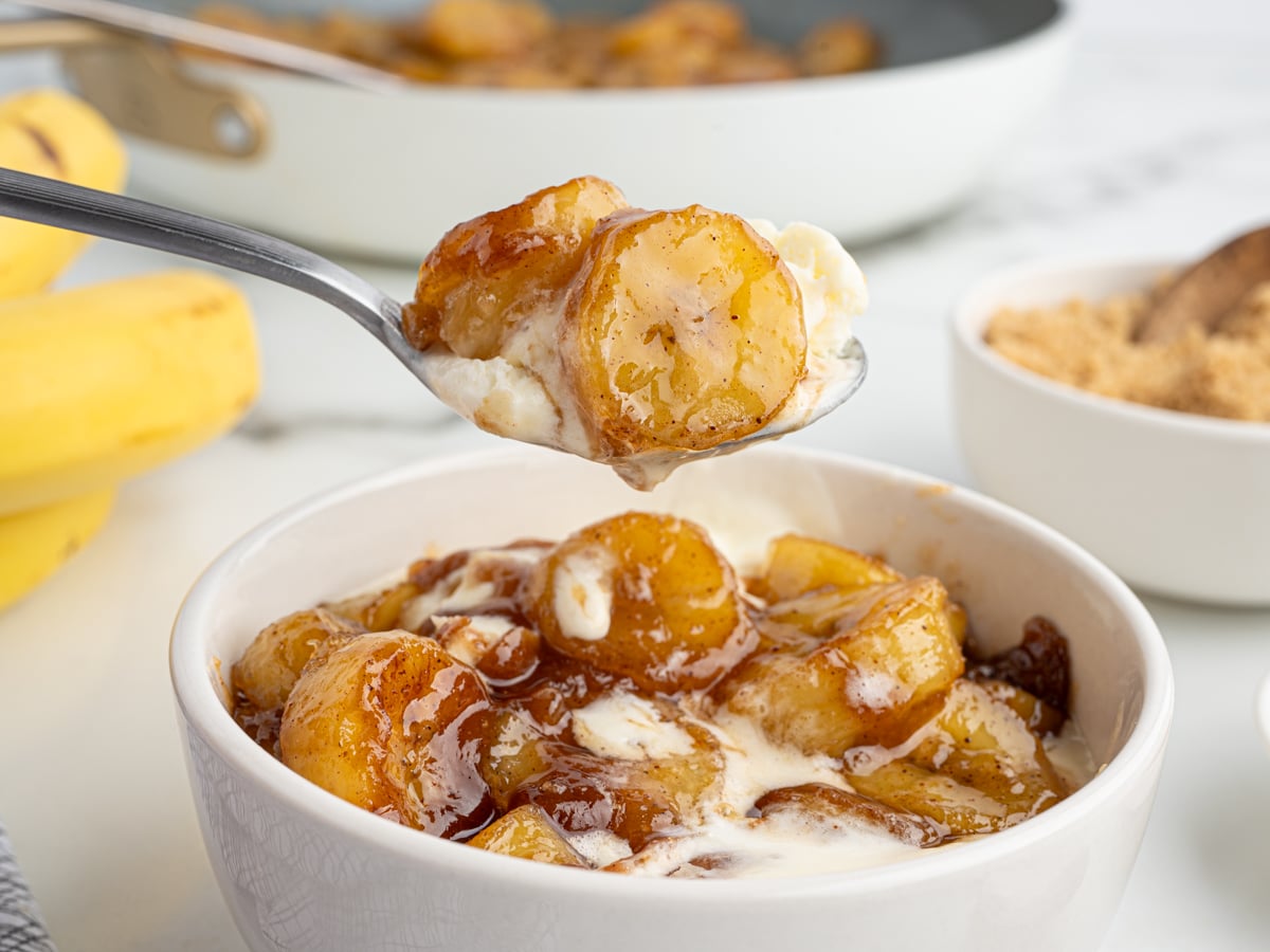 A bowl of vanilla ice cream topped with caramelized bananas; a spoon lifts a portion above the bowl. In the background, a pan and fresh bananas sit beside a bowl of brown sugar, highlighting the dessert’s rich flavors.