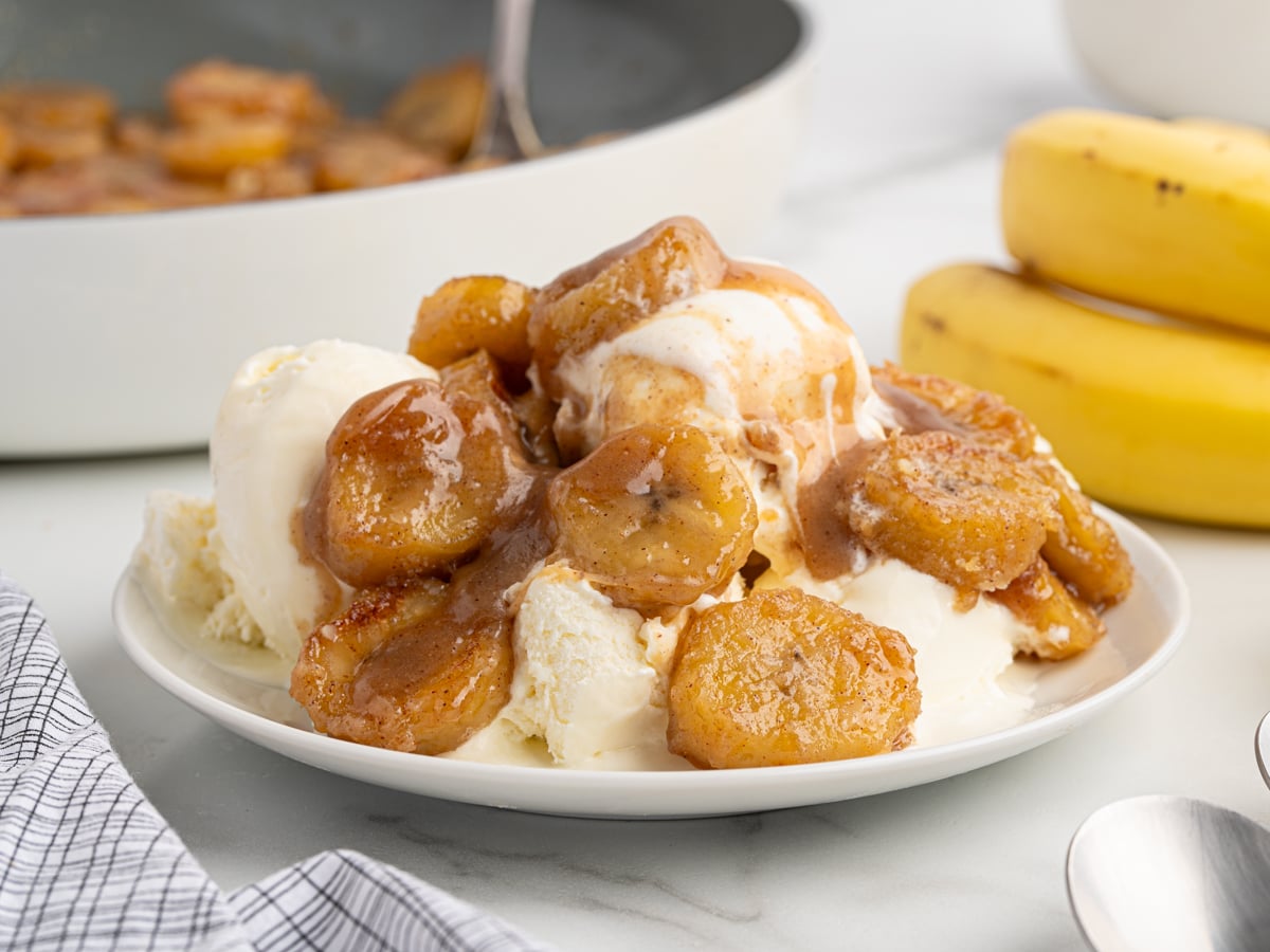 A plate of vanilla ice cream topped with caramelized bananas, with fresh bananas and a pan of more caramelized bananas in the background. A spoon and striped napkin rest beside the plate.