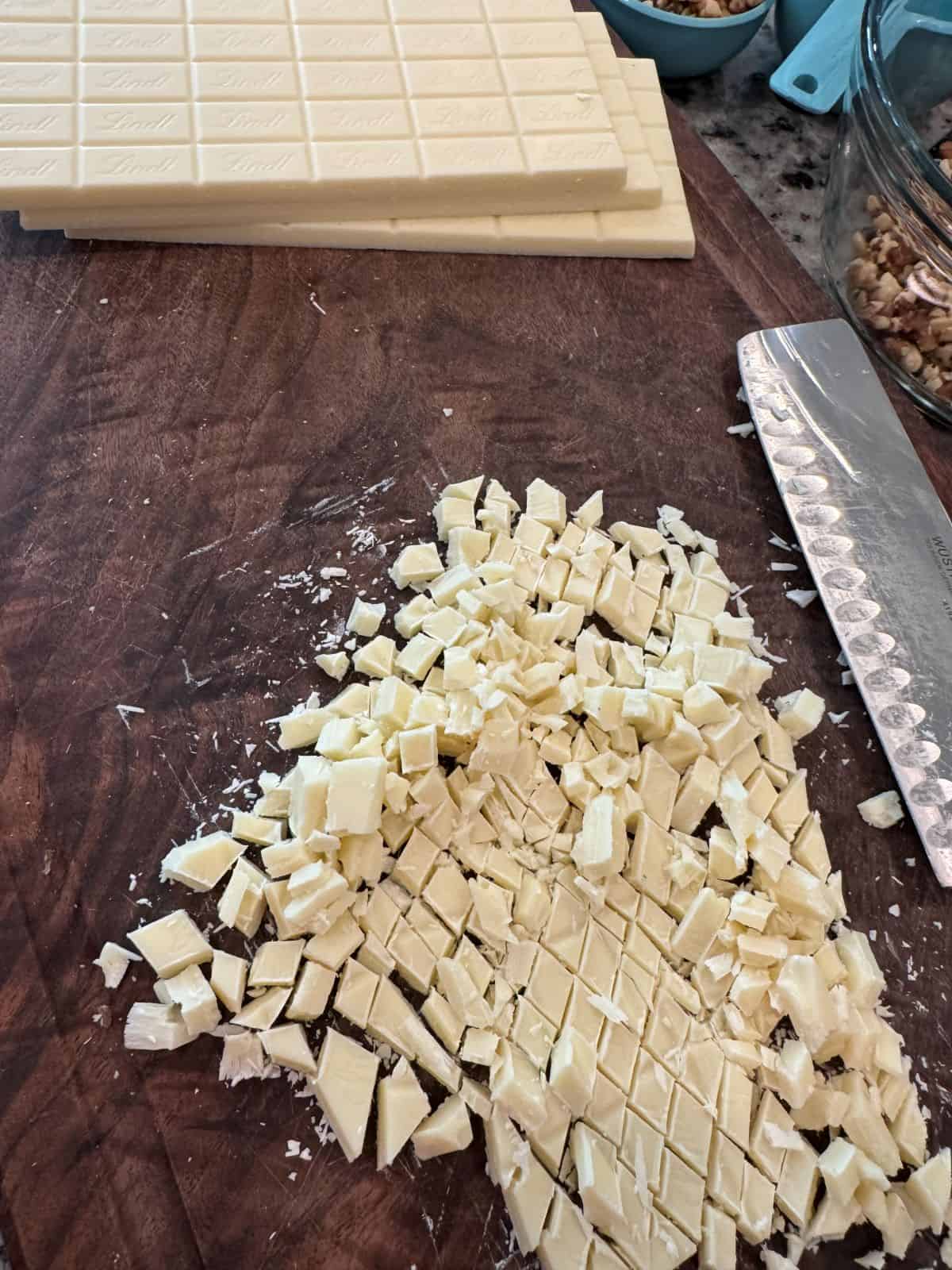Chopped white chocolate pieces on a wooden cutting board, with stacked chocolate bars and a large knife nearby—perfect for preparing homemade Maple Walnut Fudge. In the background, a glass bowl and measuring cups filled with nuts hint at the rich, nutty flavors to come.