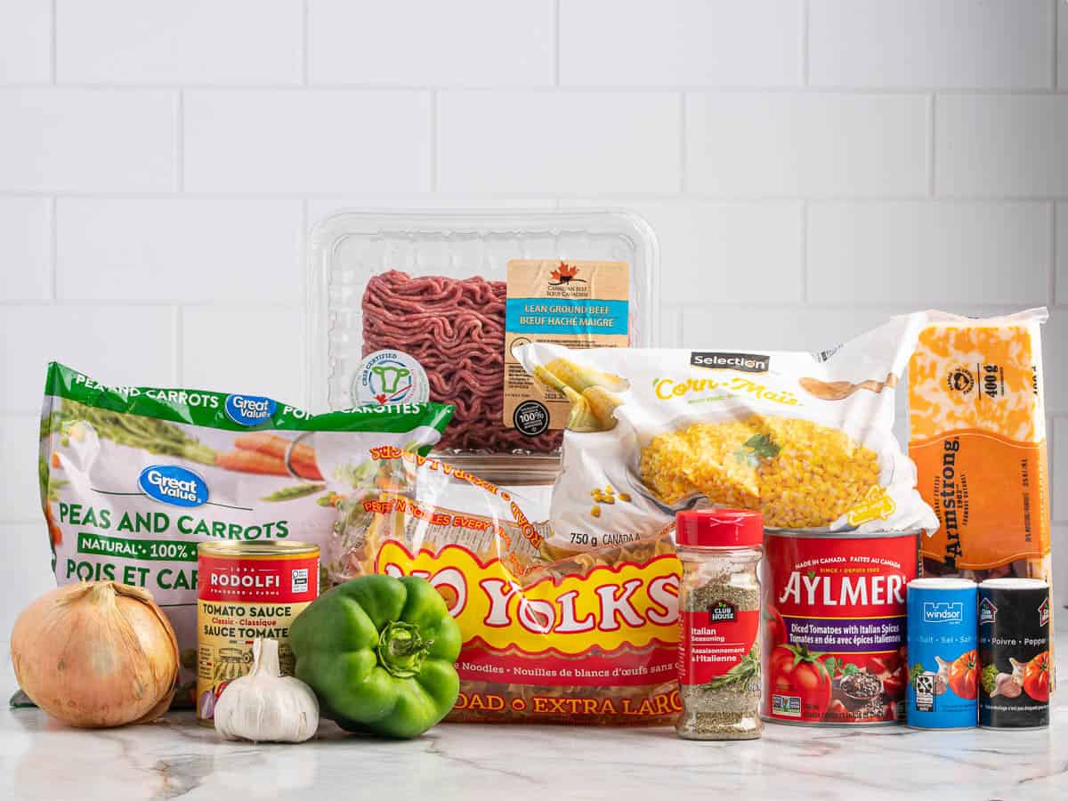 A variety of groceries on a counter—including frozen vegetables, ground beef, eggs, spices, tomato sauce, an onion, a green bell pepper, and canned goods—are displayed with a white tiled wall in the background, perfect ingredients for preparing a delicious Hamburger Casserole.