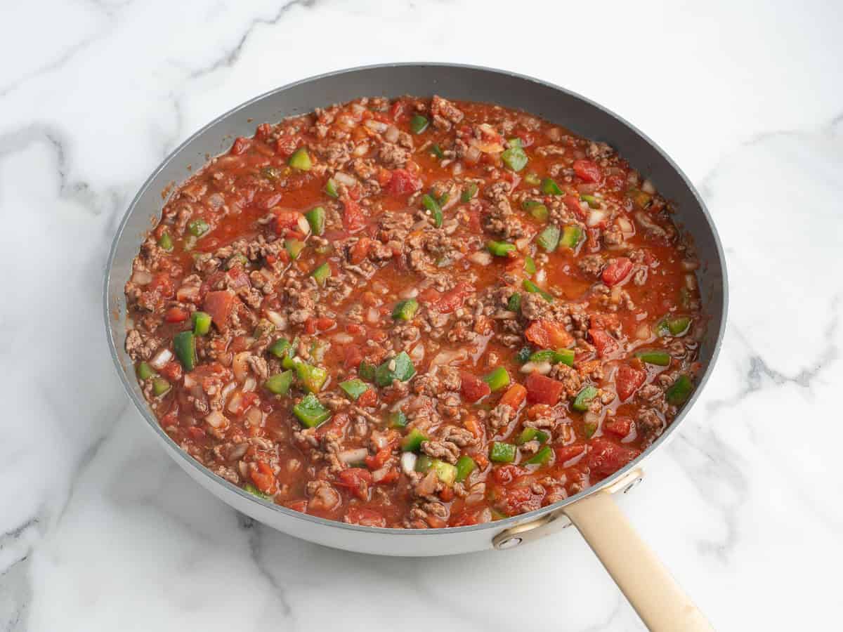 A skillet with ground beef, diced tomatoes, green bell peppers, and onions in a tomato sauce, sitting on a white marble surface—this classic dish is the perfect start to any Hamburger Casserole recipe.