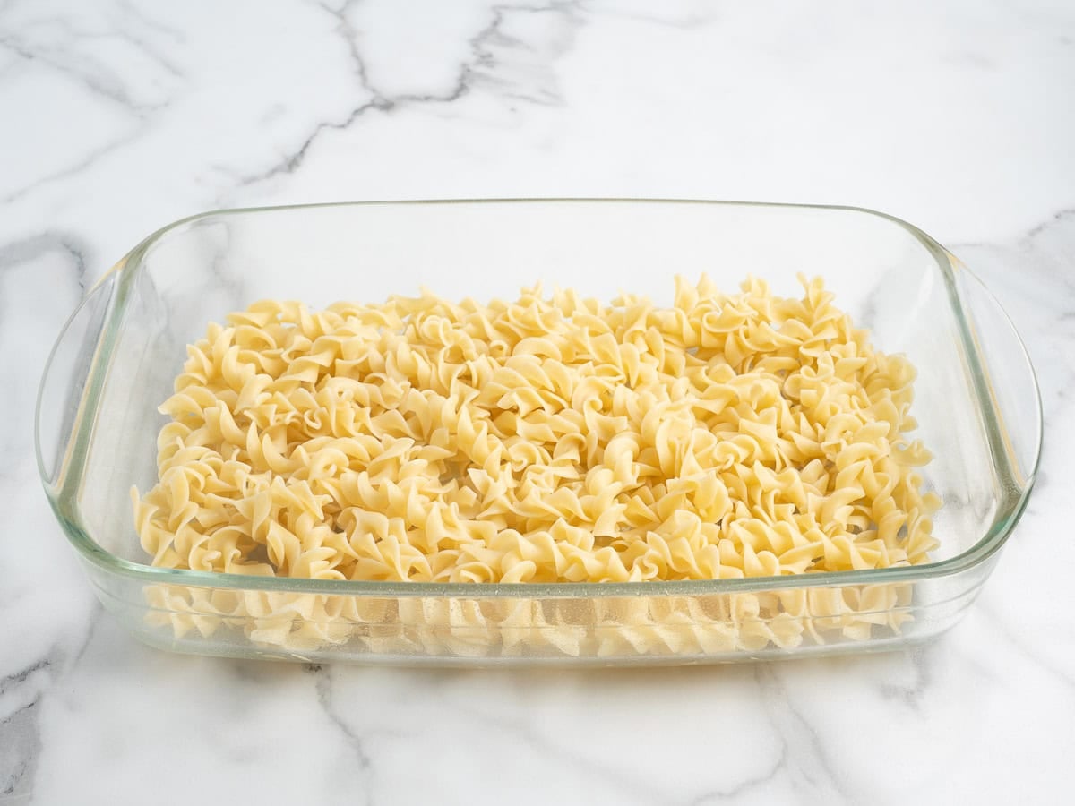 A clear glass baking dish filled with uncooked egg noodles, ready to be transformed into a delicious Hamburger Casserole, sits on a white marble surface.