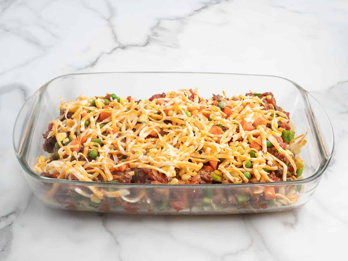 A glass baking dish filled with a layered Hamburger Casserole topped with shredded cheese, diced tomatoes, peas, and carrots, placed on a white marble surface.