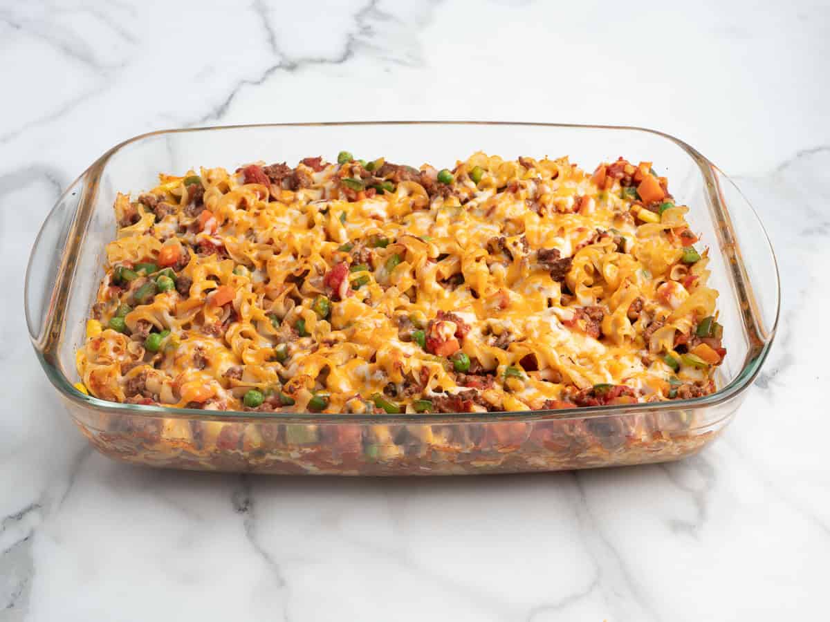 A glass baking dish filled with a baked Hamburger Casserole containing ground meat, peas, carrots, corn, tomatoes, and melted cheese, on a marble surface.