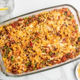 A glass baking dish filled with cheesy Hamburger Casserole made of noodles, ground meat, peas, carrots, and melted cheese sits on a white surface next to plates, a napkin, and two glasses of water with lemon.