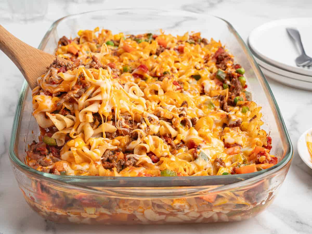 A glass baking dish filled with cheesy Hamburger Casserole, made with ground beef, diced tomatoes, and green peppers. A wooden spoon lifts a serving of this delicious Hamburger Casserole Recipe, showing melted cheese stretching invitingly. Plates and forks are in the background.
