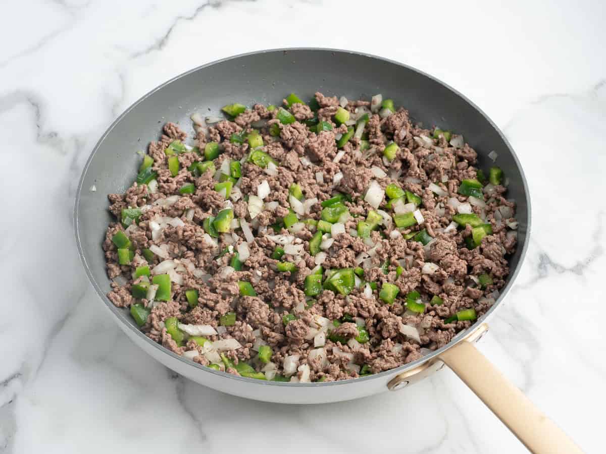 A skillet with cooked ground beef, chopped green bell peppers, and diced onions on a marble countertop—the perfect base for a classic Hamburger Casserole Recipe.