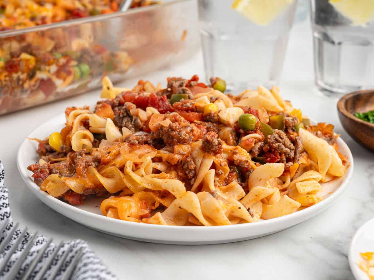 A plate of beef noodle casserole—often known as Hamburger Casserole—with ground beef, noodles, tomato sauce, corn, and green peas is served on a white plate. Two glasses of water with lemon slices are in the background, making this classic Hamburger Casserole Recipe a comforting mealtime favorite.