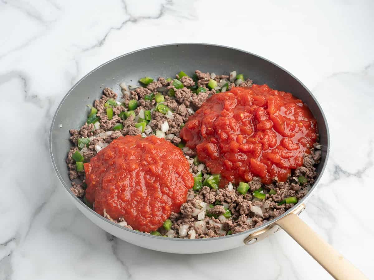 A skillet with cooked ground beef, chopped green peppers, onions, and two piles of diced tomatoes and tomato sauce on top—all the ingredients you need to start a classic Hamburger Casserole Recipe—rests on a white marble surface.