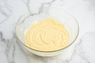A glass bowl filled with creamy, pale yellow Maple Walnut Fudge batter sits on a white marble surface.