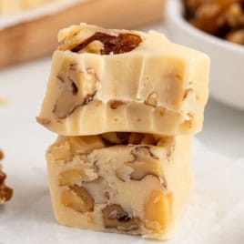 Two pieces of creamy white Maple Walnut Fudge with visible chunks of walnuts are stacked on a white surface. A bowl of walnuts is blurred in the background.