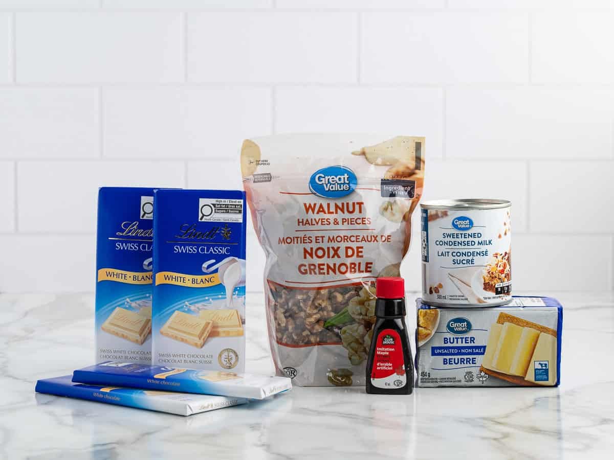 Six baking ingredients—white chocolate bars, walnuts, vanilla extract, sweetened condensed milk, and butter—are arranged on a marble counter with a white tile background, ready to be transformed into rich Maple Walnut Fudge.