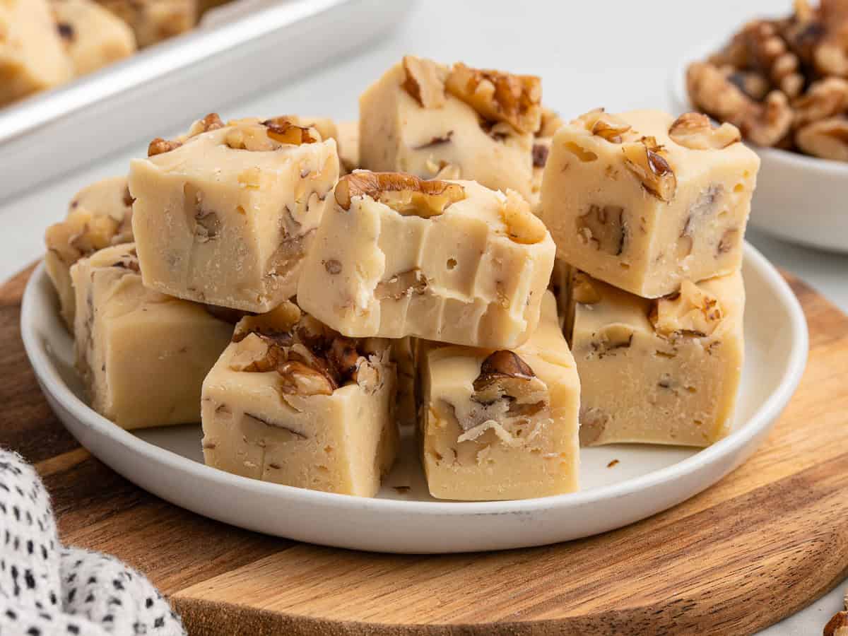 A plate stacked with creamy, nut-filled Maple Walnut Fudge squares, showcasing visible walnut pieces. The plate sits on a wooden board, with more fudge and walnuts in the background.