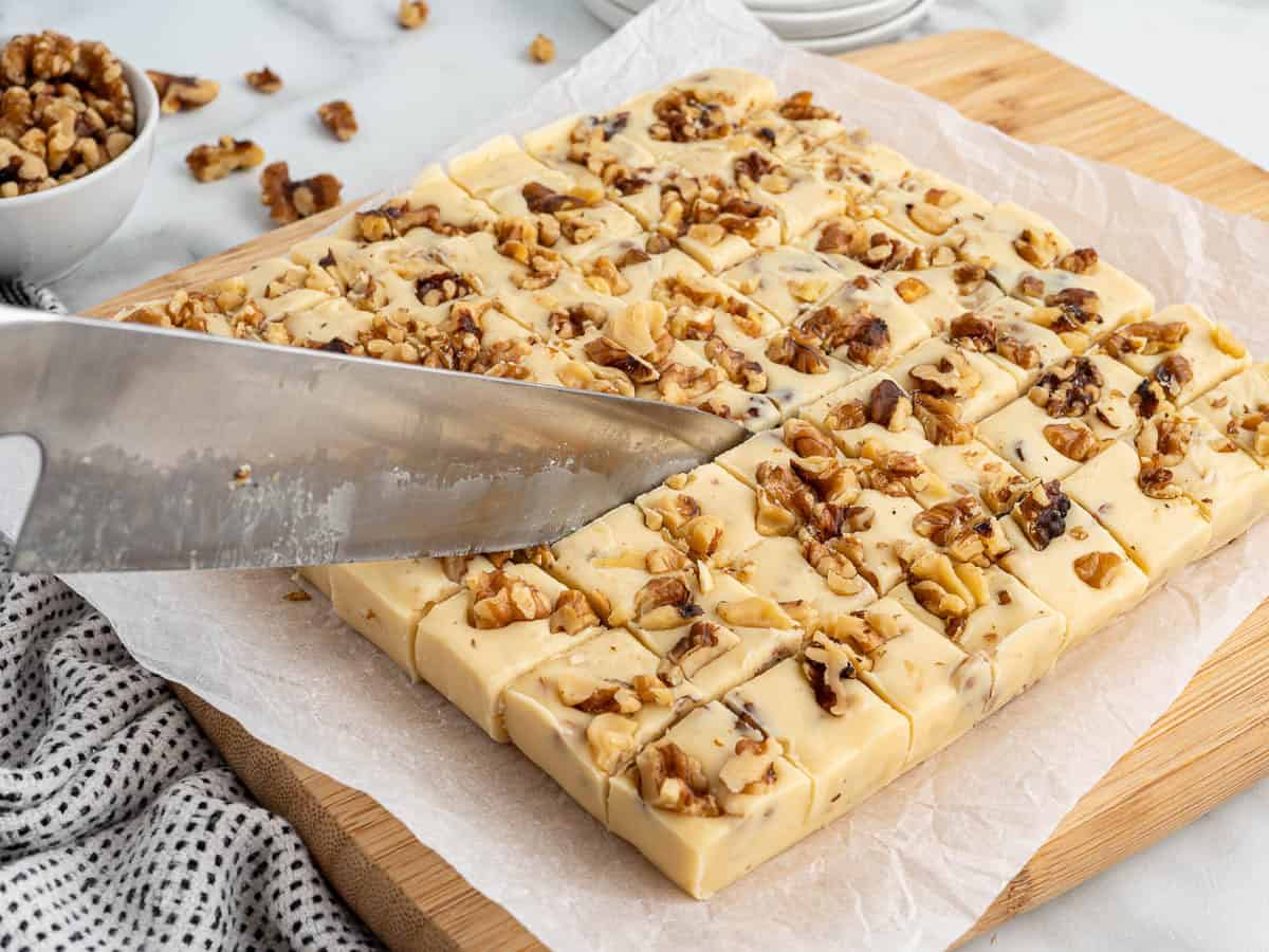 A large knife slices a slab of Maple Walnut Fudge, its creamy white surface generously topped with chopped walnuts, which is cut into small squares on parchment paper atop a wooden board. A bowl of walnuts and a striped cloth are nearby.