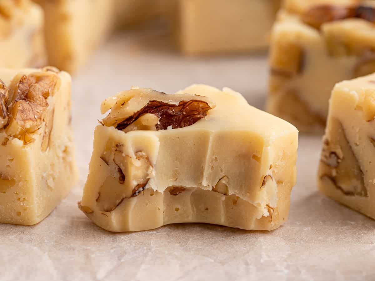 Close-up of creamy Maple Walnut Fudge squares on parchment paper, with one piece showing a bite taken out, revealing nuts and a smooth texture inside.