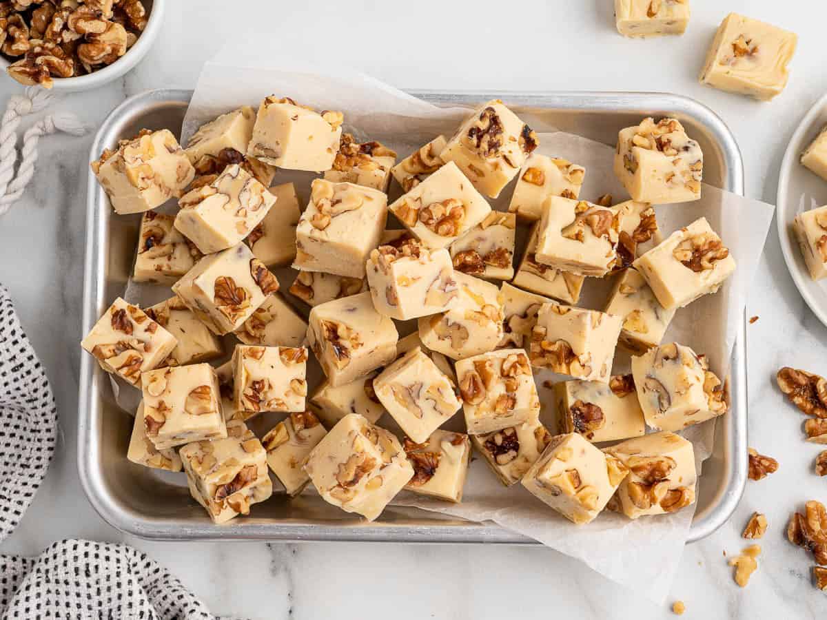 A tray filled with square pieces of creamy Maple Walnut Fudge topped with chopped walnuts, some fudge pieces slightly askew. Additional walnuts and a bowl with more Maple Walnut Fudge are visible nearby on a marble surface.