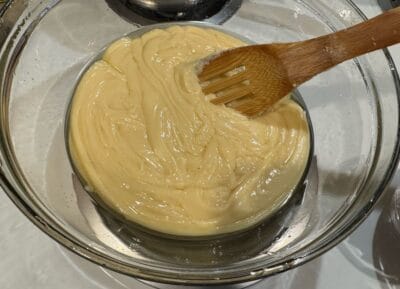 A wooden brush spreads a smooth, creamy yellow batter in a glass bowl, preparing it for baking. The mixture appears thick and glossy, reminiscent of the rich texture found in Maple Walnut Fudge, and the bowl sits on a white countertop.