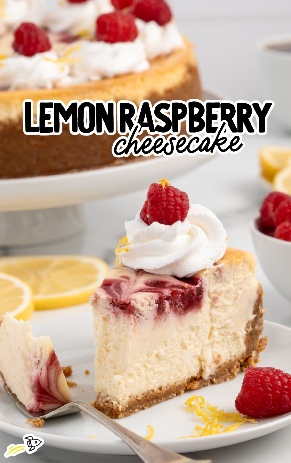 A slice of lemon raspberry cheesecake on a white plate, showing a creamy interior with a vibrant raspberry swirl on top. The slice is garnished with whipped cream, a fresh raspberry, and lemon zest, with the whole cheesecake and fresh raspberries visible in the background.
