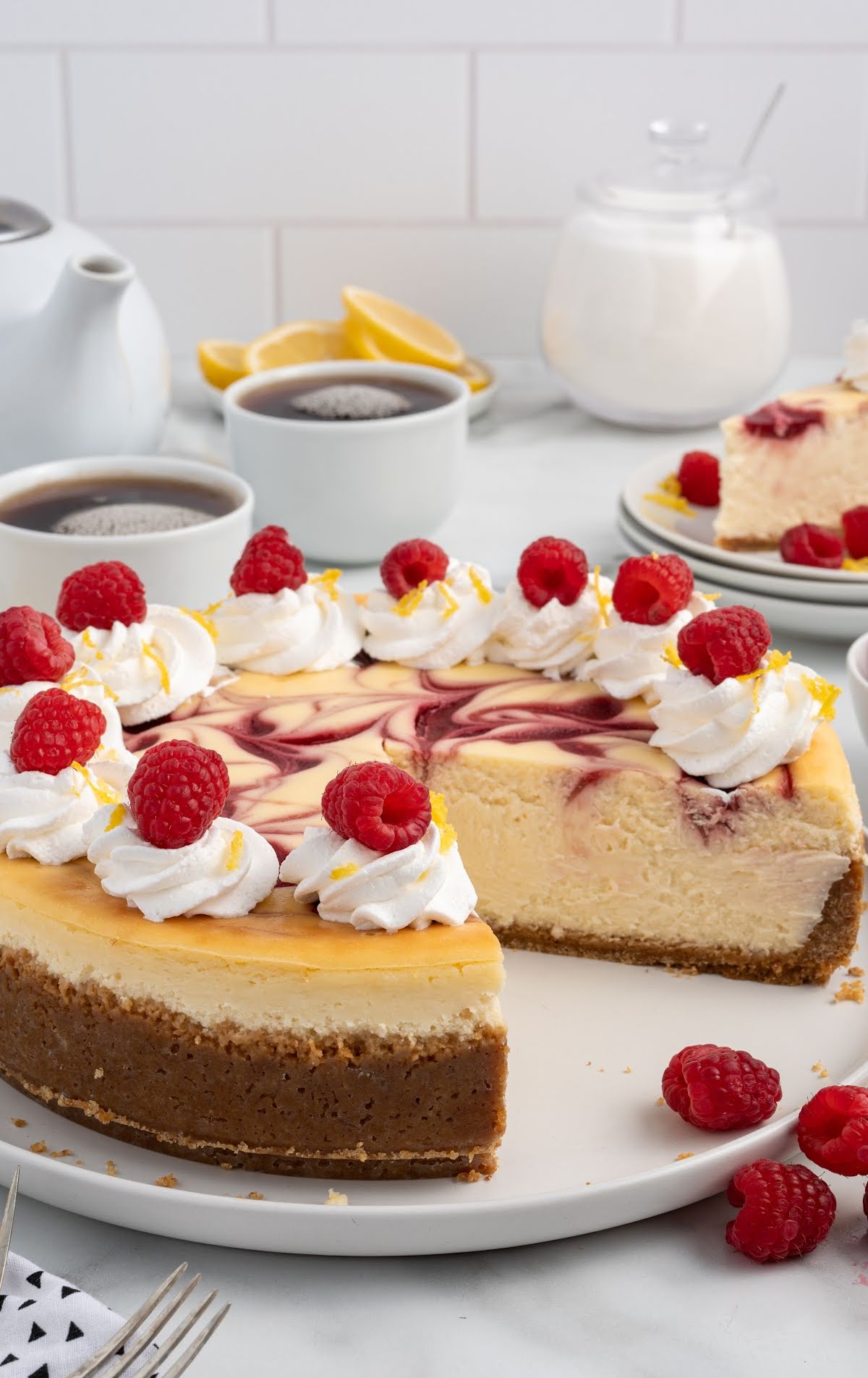Slice of lemon raspberry cheesecake on a white plate, showing a creamy interior with a raspberry swirl on top, garnished with whipped cream, a fresh raspberry, and lemon zest. The remaining cheesecake sits on a cake stand in the background, with lemon slices and fresh raspberries around the plate.