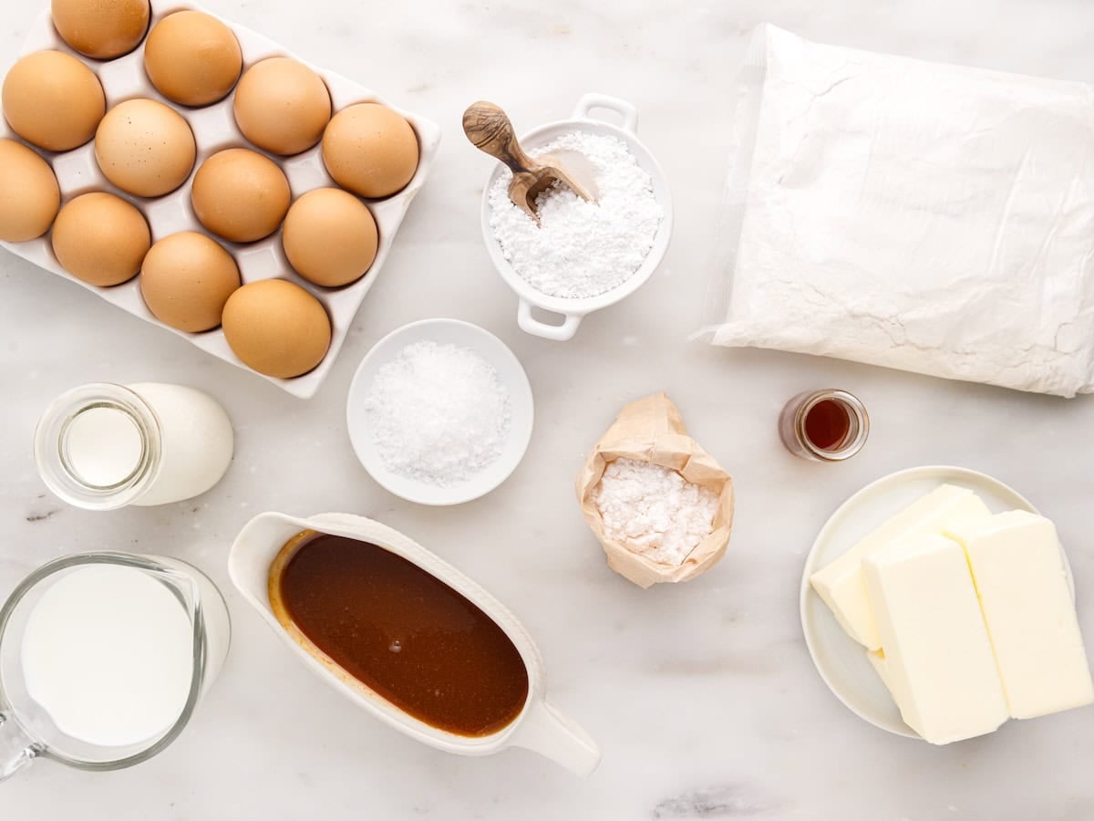 Baking ingredients on a white surface: eggs in a carton, a block of butter, milk, cream, flour, powdered sugar, vanilla extract, brown sugar syrup, and a small bowl of salt—all you need to create the perfect salted caramel drip cake.