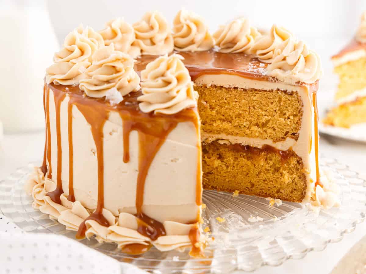A salted caramel drip cake with creamy frosting, topped with decorative swirls, sits on a glass platter. A slice is removed, revealing the moist, golden interior beneath the luscious caramel drizzle.