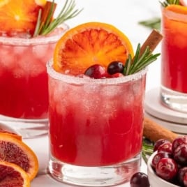 Three glasses of cranberry Christmas cocktail with sugared rims, garnished with orange slices, cranberries, and rosemary.