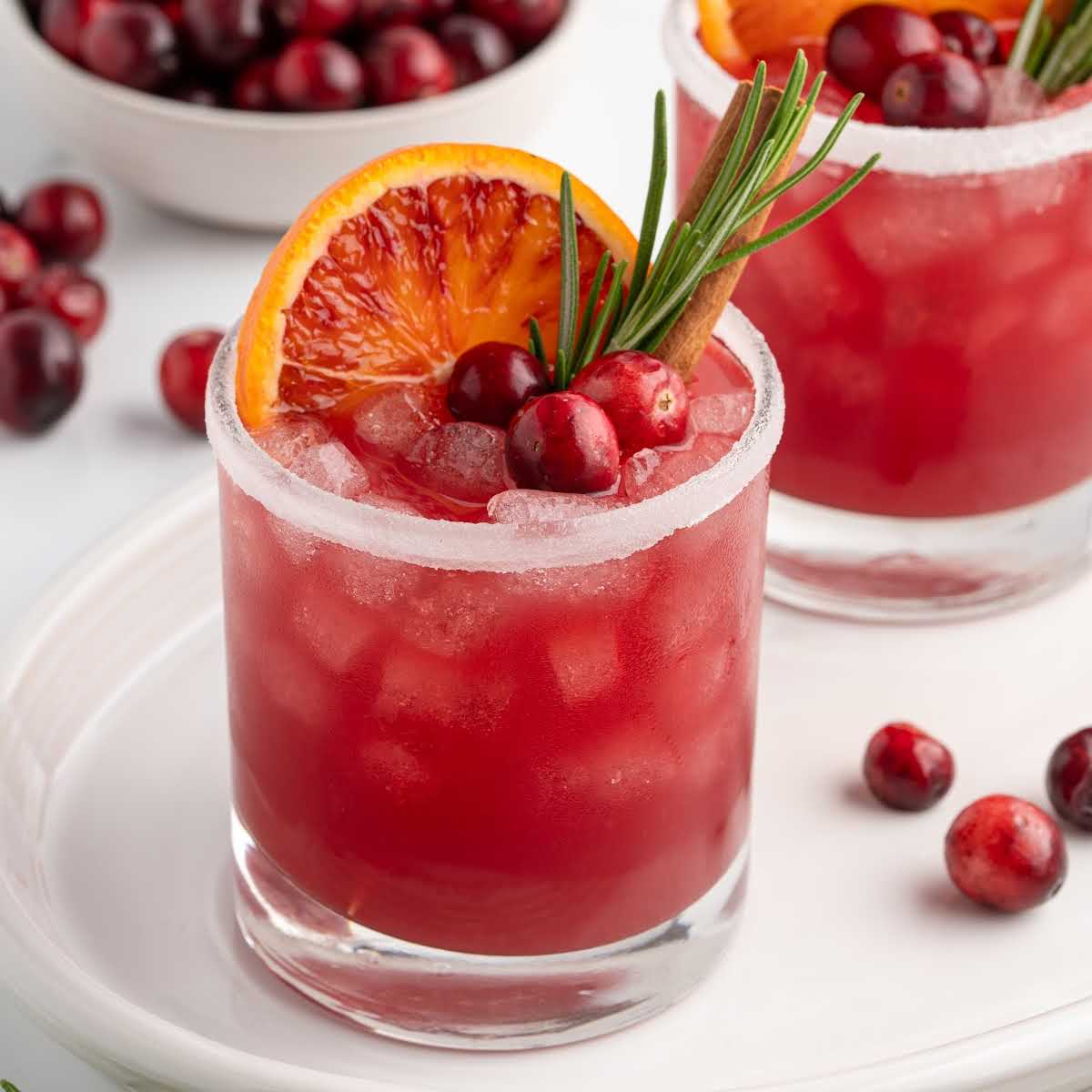 Close-up of cranberry Christmas cocktail in a sugared glass garnished with cranberries, rosemary, and an orange slice.