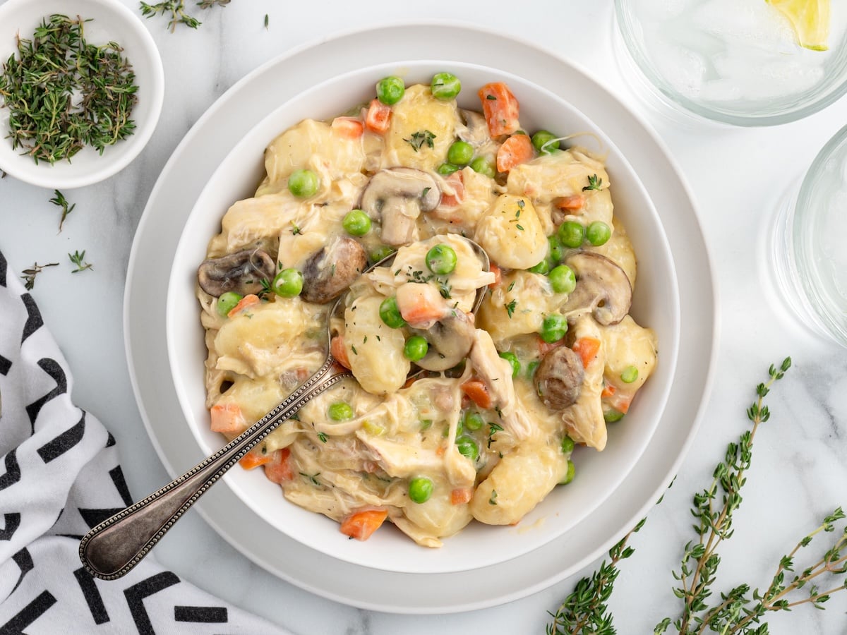 A bowl of creamy gnocchi chicken pot pie with peas, carrots, mushrooms, and herbs, served with a silver spoon. Fresh thyme and a glass of water with lemon rest nearby on a white marble surface.
