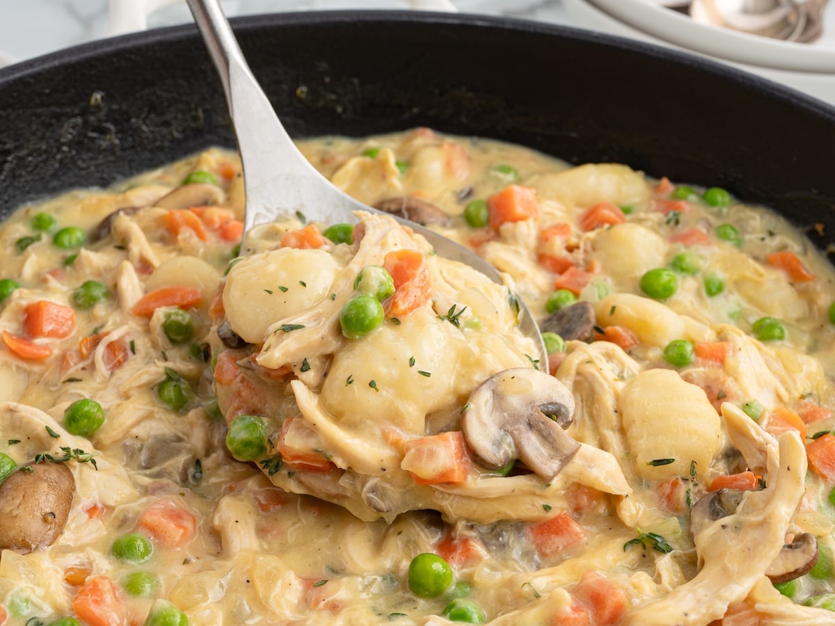 A spoon lifts creamy Gnocchi Chicken Pot Pie&mdash;packed with tender chicken, gnocchi, peas, carrots, and mushrooms&mdash;from a black skillet, highlighting the hearty texture of the dish.