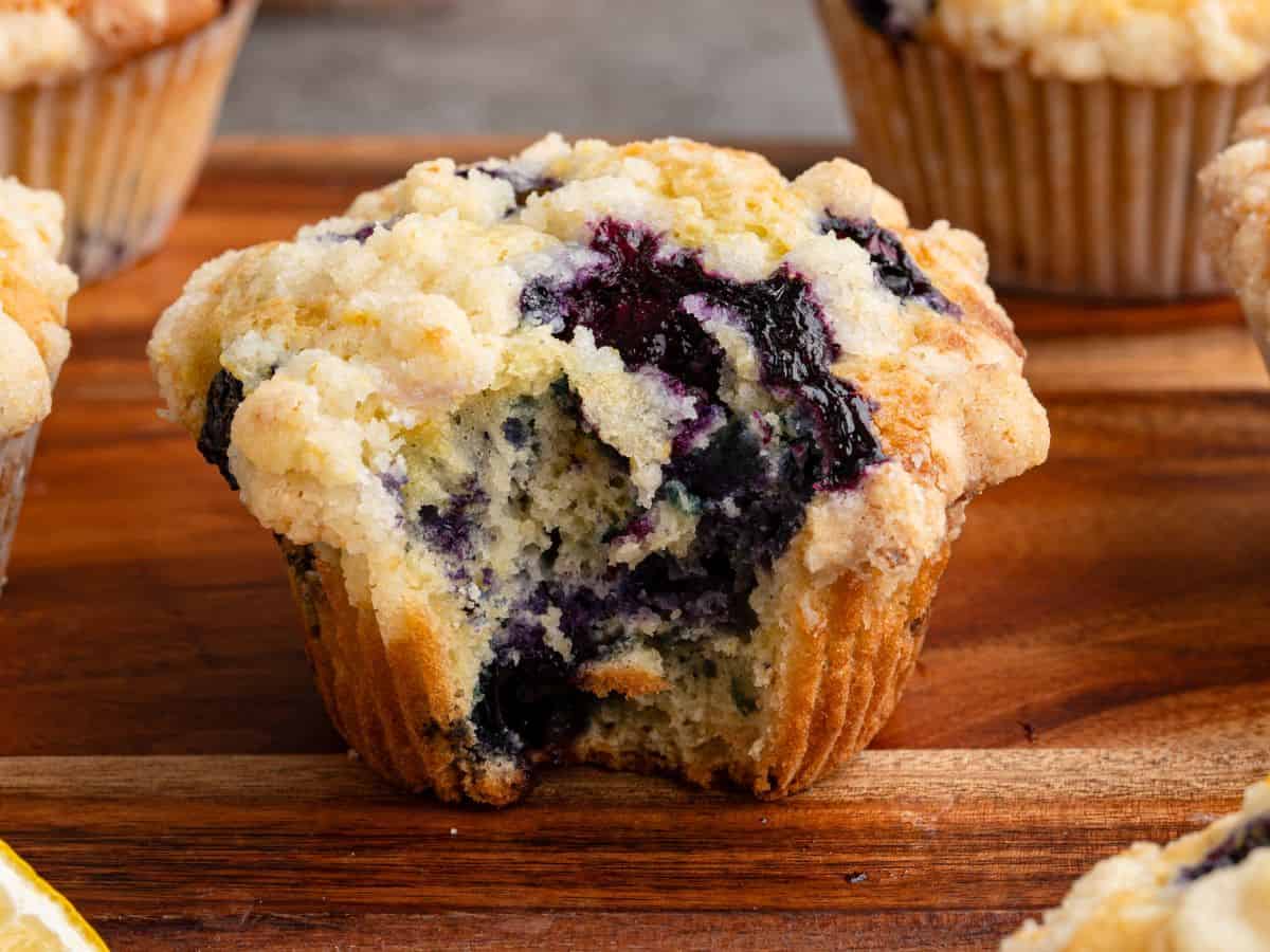 A Lemon Blueberry Muffin with a streusel topping sits on a wooden board. The muffin has a large bite taken out, revealing its soft, blueberry-filled interior with a hint of citrus. Other Lemon Blueberry Muffins are visible in the background.