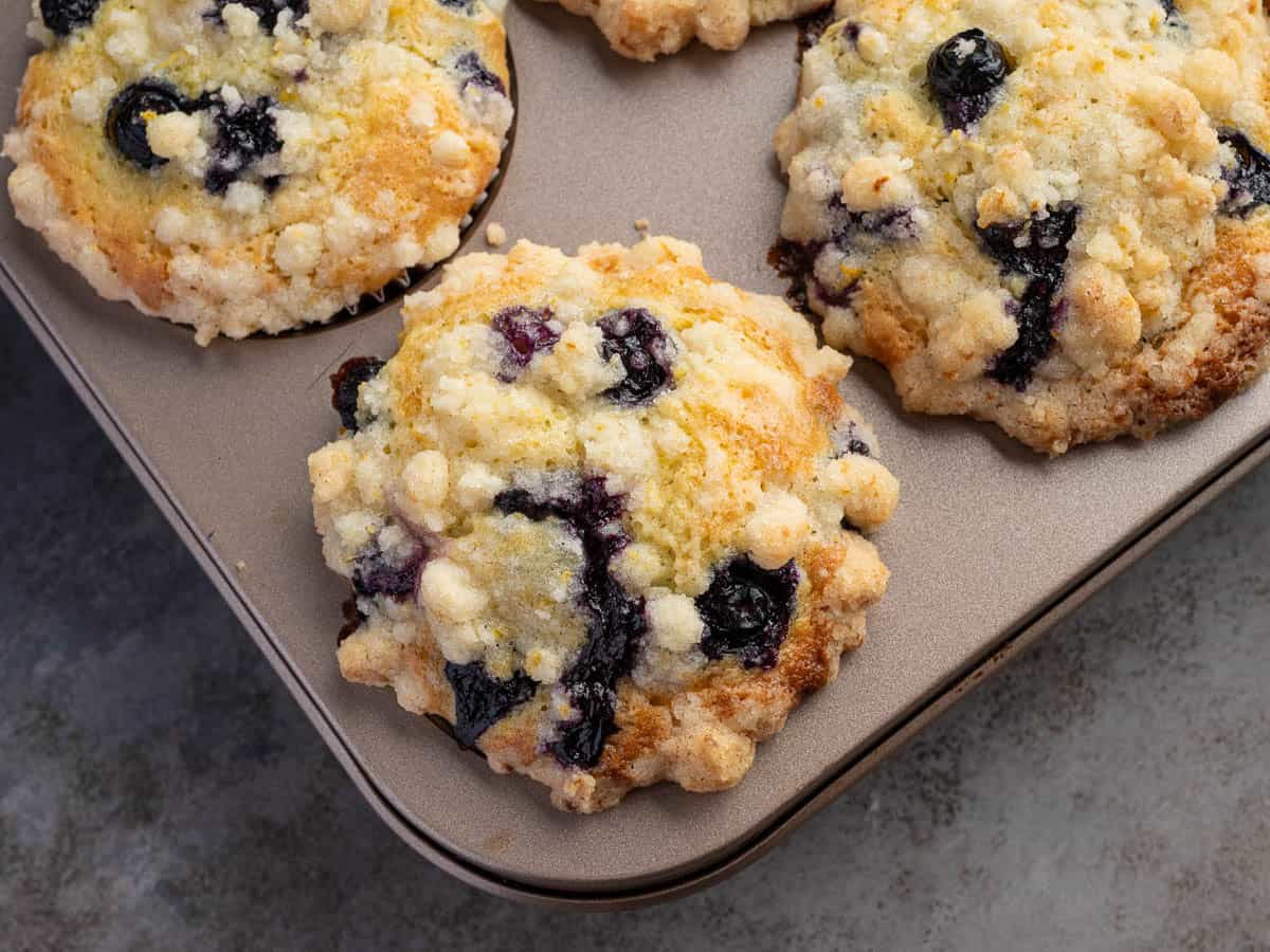 Three freshly baked Lemon Blueberry Muffins with crumbly topping sit in a muffin tin on a gray surface.