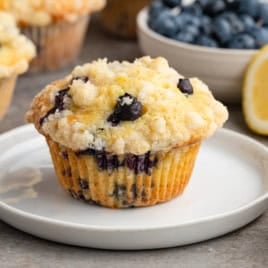 A Lemon Blueberry Muffin with crumb topping sits on a white plate. In the background are fresh blueberries and a sliced lemon, suggesting the muffins’ bright, zesty flavors.