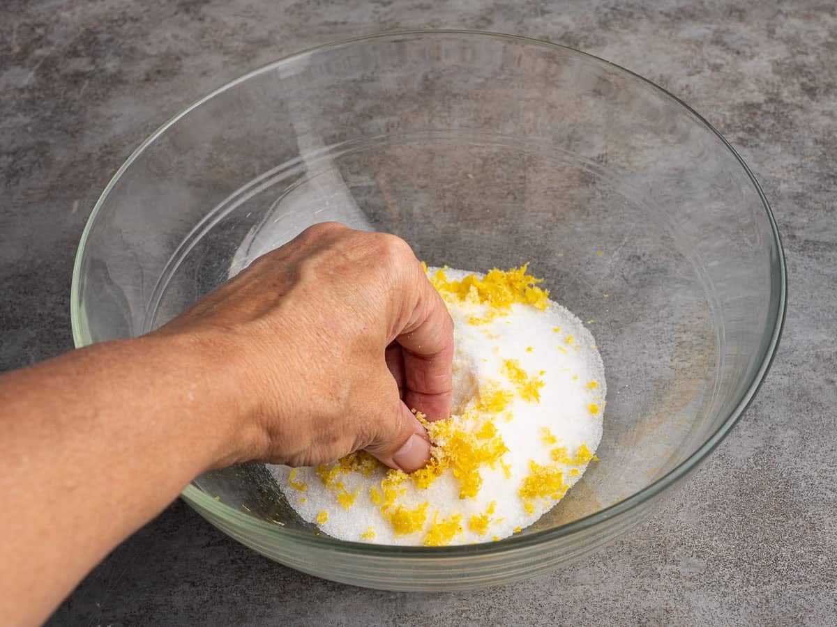 A hand rubs lemon zest into granulated sugar in a clear glass bowl on a gray countertop, preparing the fragrant base for homemade Lemon Blueberry Muffins.
