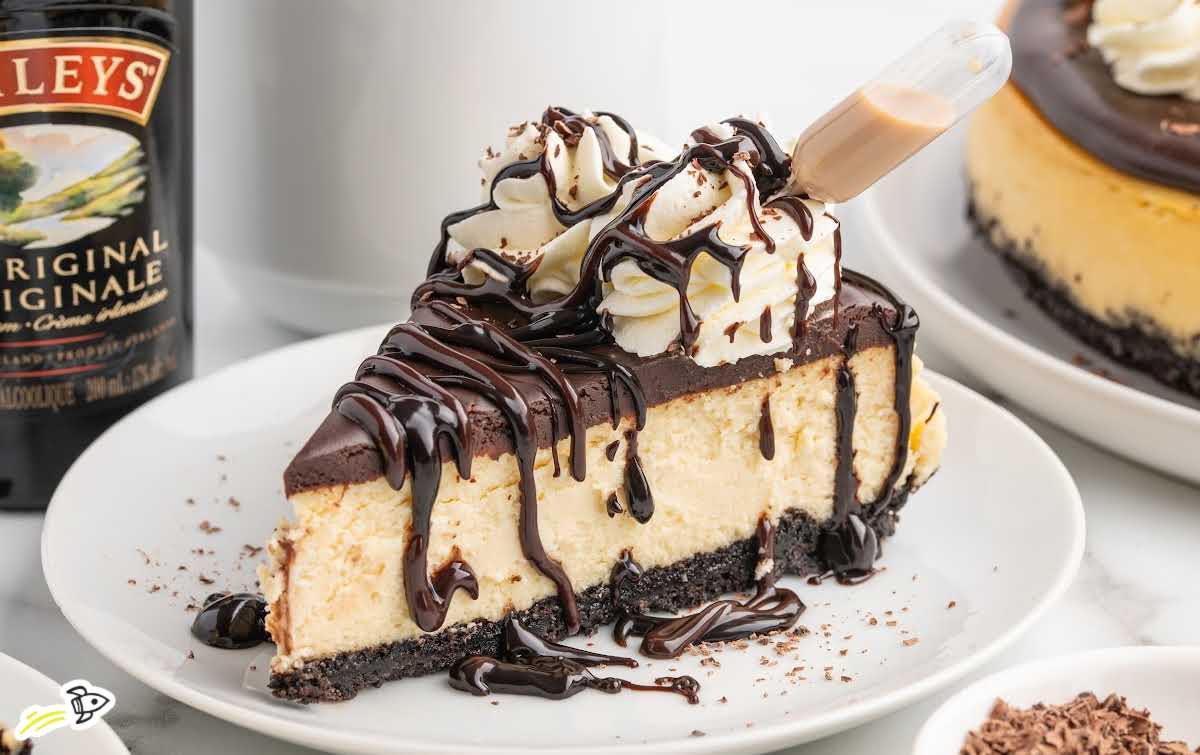 Slice of Baileys cheesecake lifted from the whole cake, showing creamy layers and Oreo crust beneath smooth ganache.