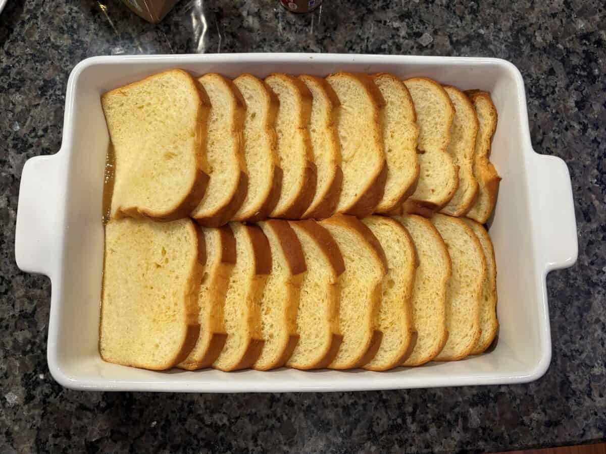 A white rectangular baking dish filled with two rows of neatly arranged sliced bread, ready to be transformed into delicious Baked French Toast, sits on a granite countertop.