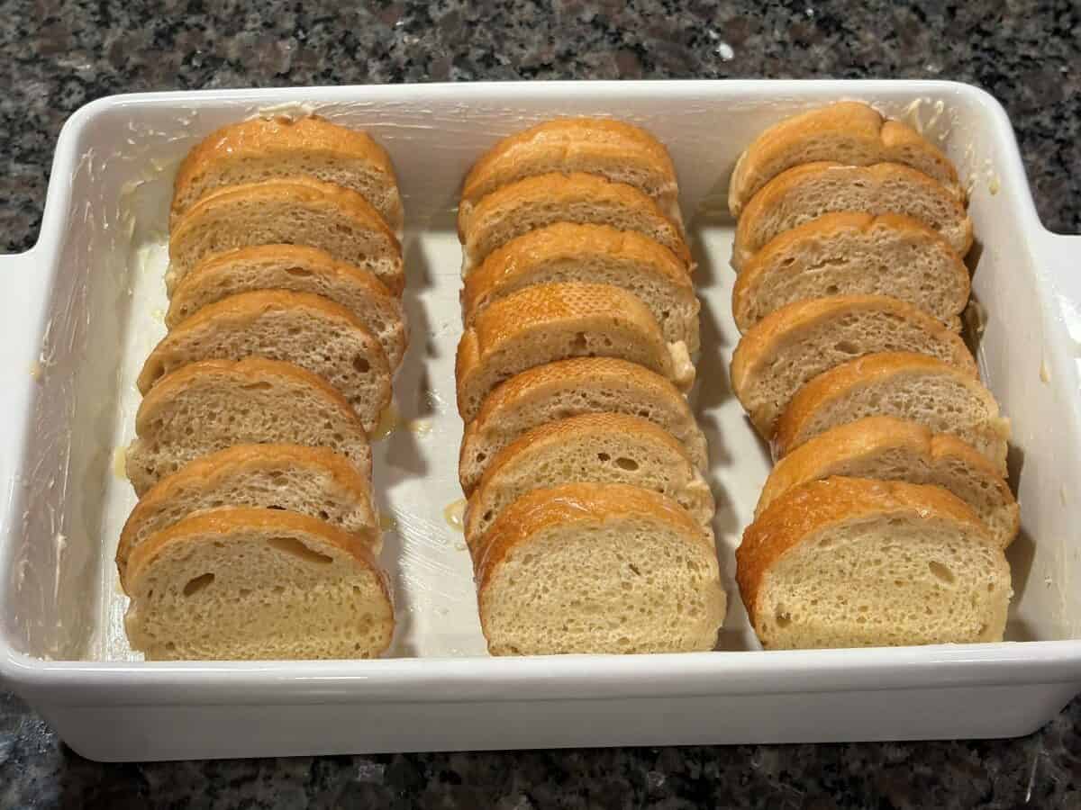 A white baking dish contains two neat rows of sliced bread arranged upright on a dark granite countertop, ready to be transformed into a delicious Baked French Toast.