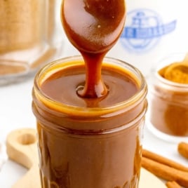 Thick brown sugar caramel sauce pouring from a spoon into a mason jar with cinnamon sticks in the background