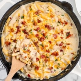 Skillet filled with crack chicken penne topped with bacon, cheddar cheese, and fresh parsley on a marble countertop.