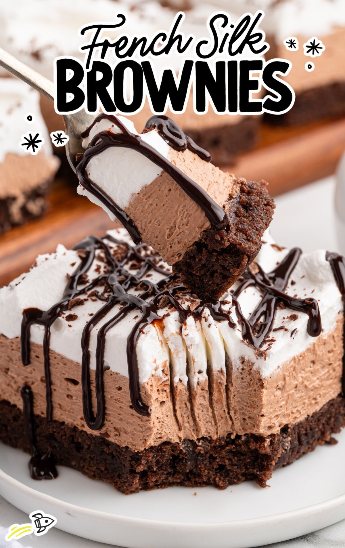A close-up of a French silk brownie on a white plate showing three layers — a chewy chocolate brownie base, a fluffy chocolate mousse middle, and a whipped cream topping drizzled with chocolate sauce. A fork lifts a bite to reveal the creamy texture.