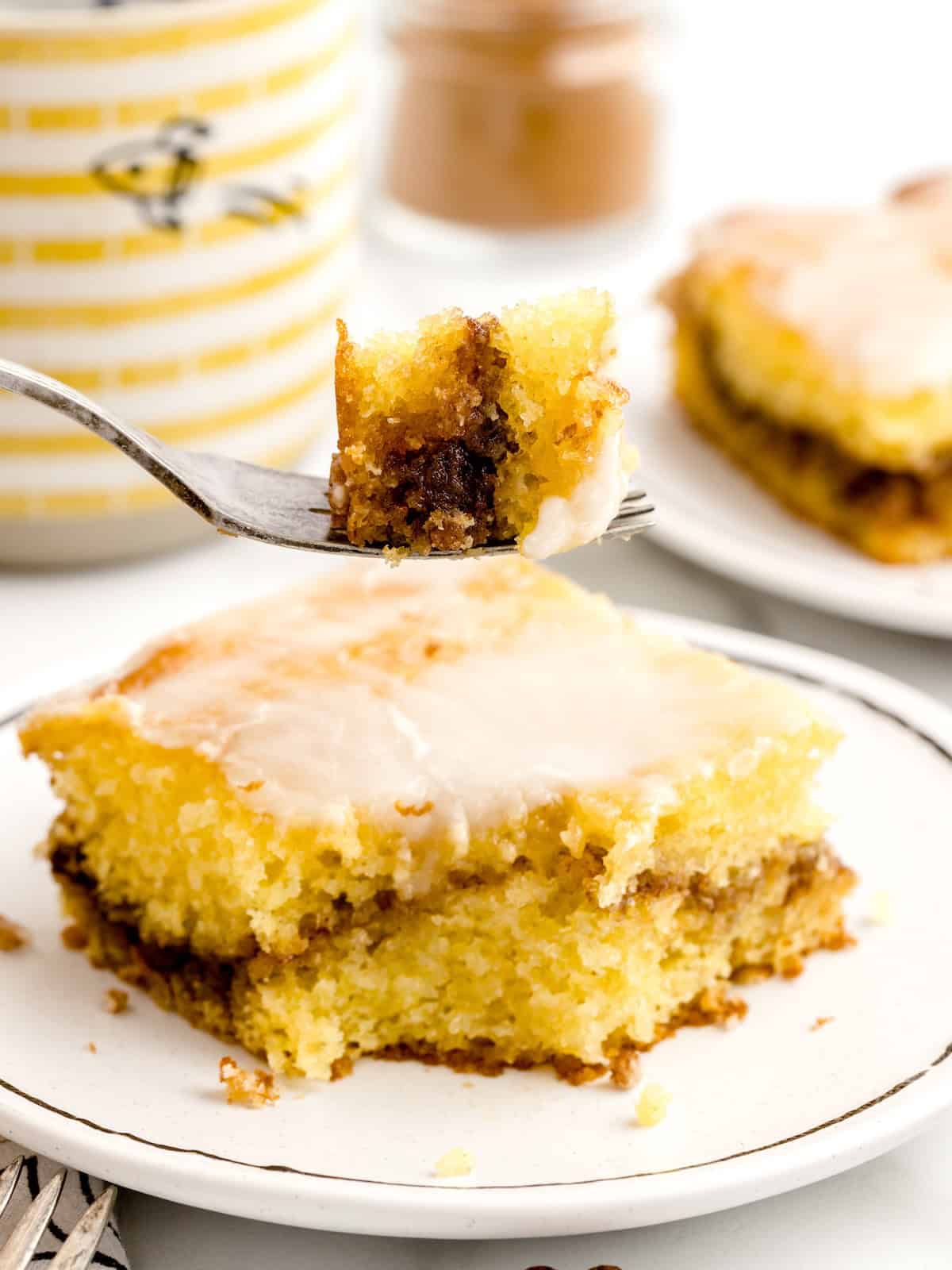 A fork holds a bite of glazed cinnamon honey bun cake above a serving on a plate.
