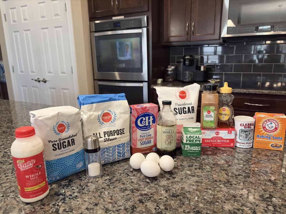 Ingredients for a Honey Bun Cake arranged on a kitchen counter, including milk, granulated sugar, flour, light brown sugar, eggs, vanilla extract, powdered sugar, buttermilk, butter, cinnamon, honey, baking powder, and baking soda.