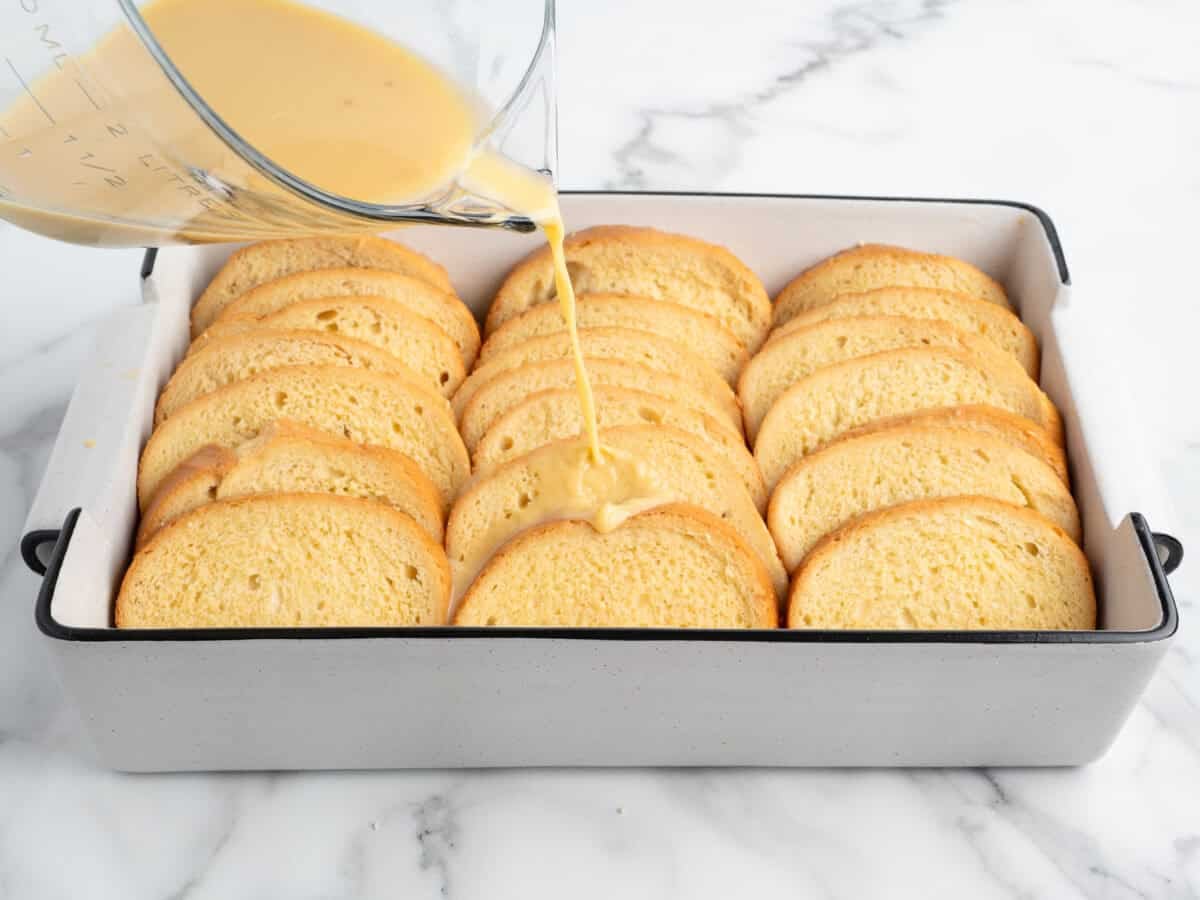 A baking dish with slices of bread arranged neatly, while a creamy liquid is being poured over them from a measuring cup on a marble countertop, prepares the perfect base for delicious Baked French Toast.