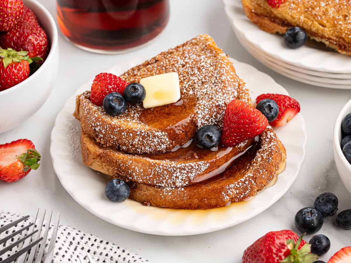 Three slices of baked French toast topped with powdered sugar, butter, syrup, strawberries, and blueberries are served on a white plate. Fresh berries are scattered around, with a fork and a bowl of strawberries nearby.