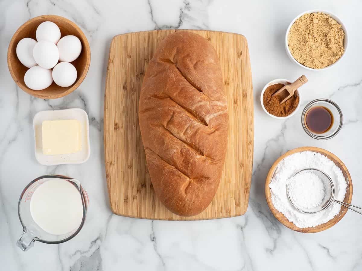 A loaf of bread on a wooden board, surrounded by eggs, butter, milk, brown sugar, cinnamon, vanilla, and powdered sugar on a marble surface, perfect ingredients for preparing delicious Baked French Toast.
