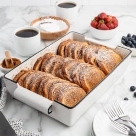 A white baking dish filled with powdered sugar-dusted Baked French Toast slices sits on a marble table, surrounded by coffee cups, a bowl of strawberries, blueberries, powdered sugar, cinnamon, and forks.