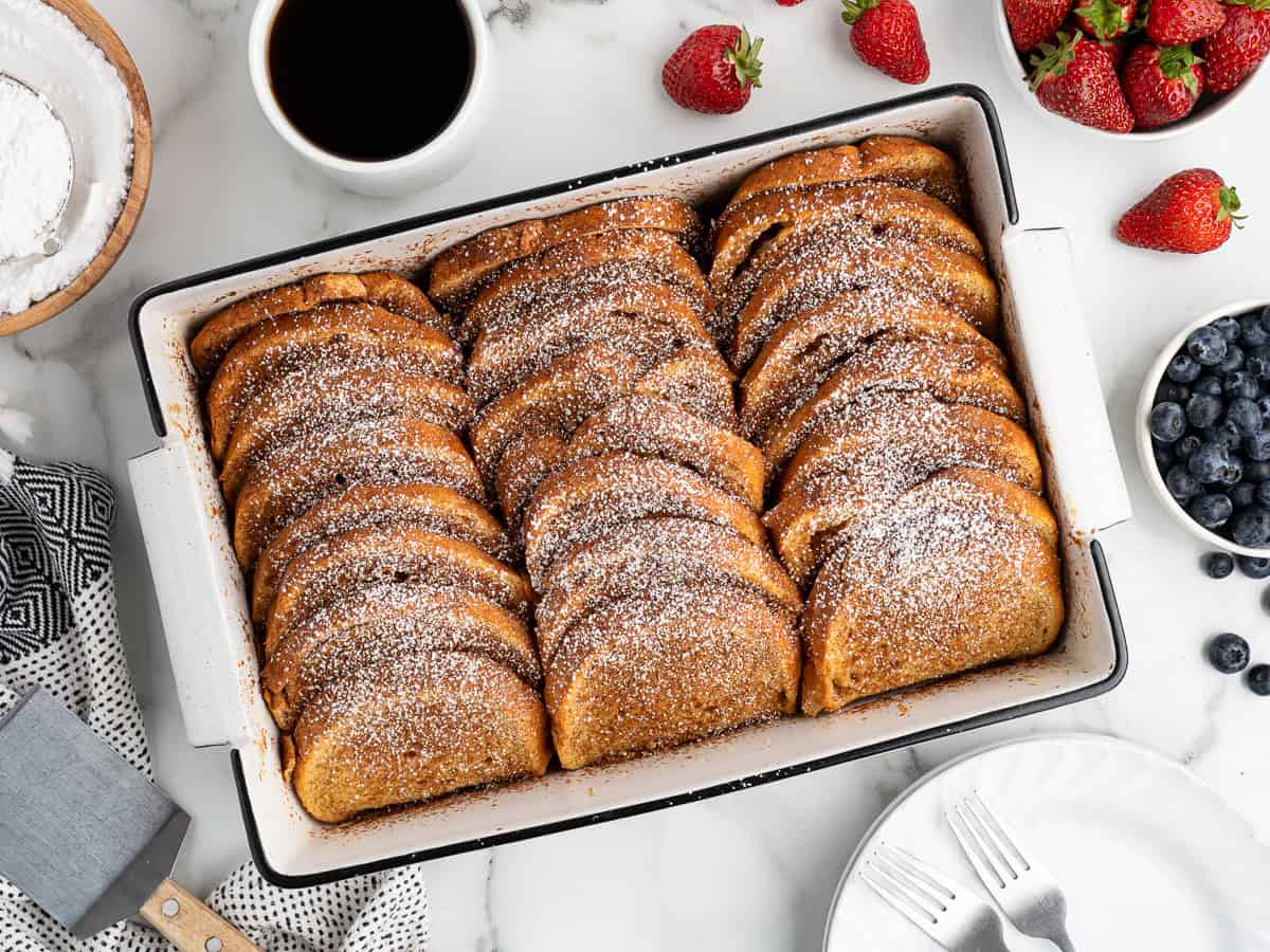 A baking dish filled with rows of golden Baked French Toast slices dusted with powdered sugar, surrounded by strawberries, blueberries, a cup of coffee, and plates on a marble surface.