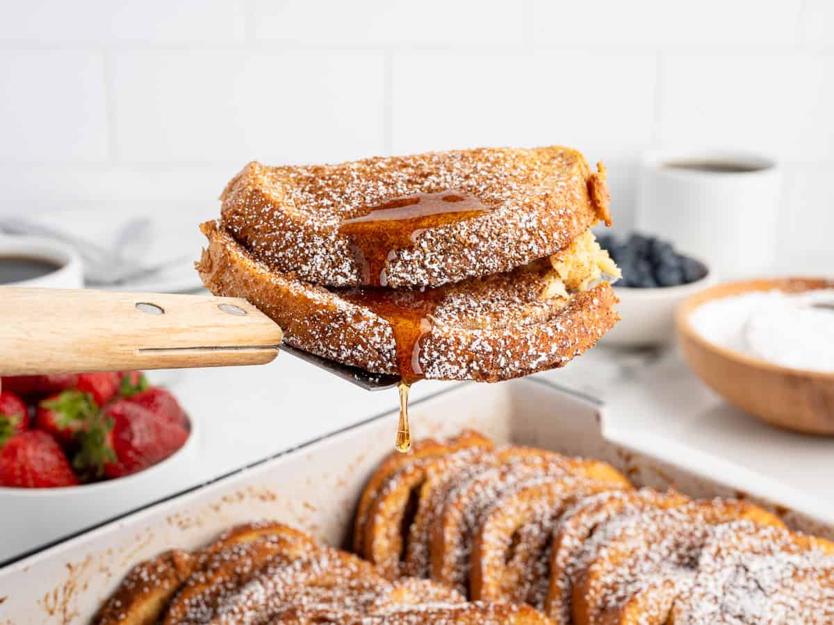 A spatula holds two slices of baked French toast dusted with powdered sugar and drizzled with syrup. More French toast, strawberries, blueberries, and coffee are in the background.
