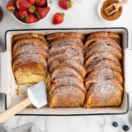 A baking dish of golden Baked French Toast slices dusted with powdered sugar, with one slice removed by a spatula. Fresh strawberries, blueberries, cinnamon, and syrup surround the dish on a marble surface.