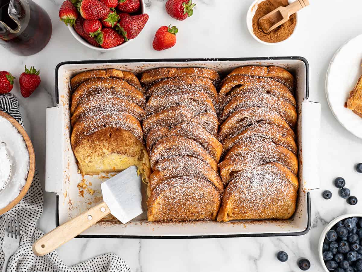 A baking dish of golden Baked French Toast slices dusted with powdered sugar, with one slice removed by a spatula. Fresh strawberries, blueberries, cinnamon, and syrup surround the dish on a marble surface.