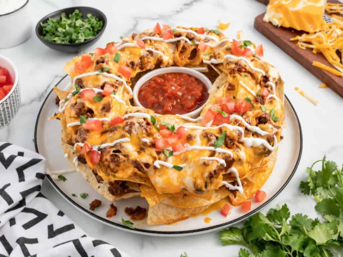 A round platter of Bundt Pan Nachos topped with melted cheese, ground beef, diced tomatoes, cilantro, and sour cream, arranged around a bowl of salsa. Shredded cheese, chopped herbs, and tomatoes are nearby.